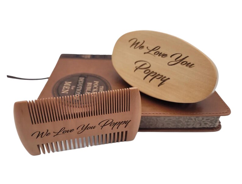 May include: A wooden beard comb and brush set with the engraved message "We Love You Poppy." The set rests on a brown leather-bound book. The comb is rectangular, and the brush is oval-shaped.
