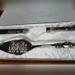 May include: A silver cake knife and server set in a white box. The server is engraved with "Mr & Mrs Ledford May 9, 2020". The knife is engraved with "Justice & Jennir".