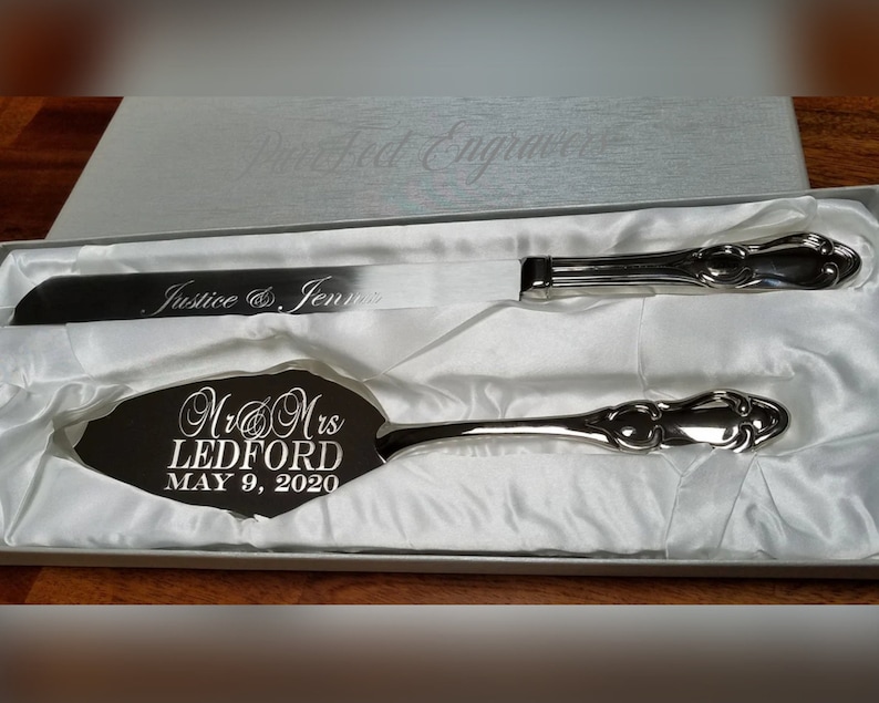 May include: A silver cake knife and server set in a white box. The server is engraved with "Mr & Mrs Ledford May 9, 2020". The knife is engraved with "Justice & Jennir".