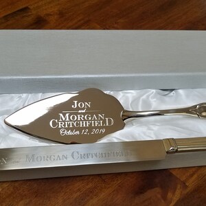 May include: Silver wedding cake knife and server set in a white box with a satin lining. The knife and server are engraved with "Jon and Morgan Critchfield, October 12, 2019".