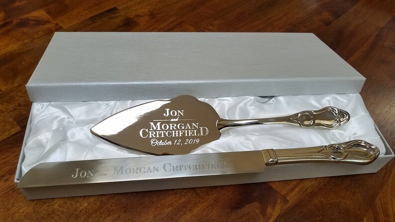 May include: Silver wedding cake knife and server set in a white box with a satin lining. The knife and server are engraved with "Jon and Morgan Critchfield, October 12, 2019".