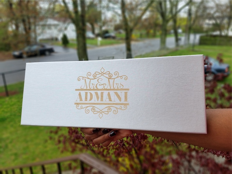 May include: A white box with a gold inscription that reads "Mr. & Mrs. ADMANI" with a decorative design.
