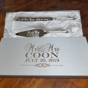 May include: A white box with a silver satin lining contains a silver cake knife and server set. The box is engraved with "Mr & Mrs COON JULY 20, 2019". The knife and server are also engraved with the same text.
