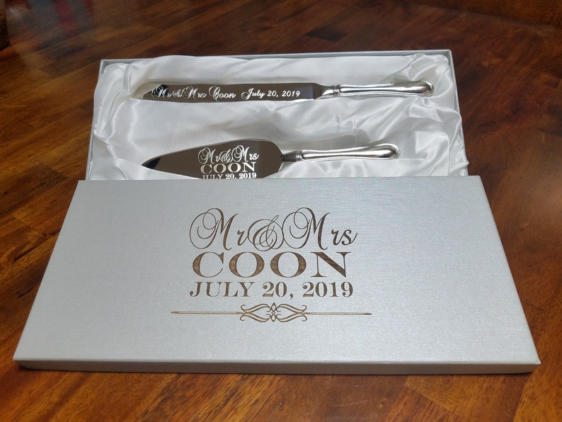 May include: A white box with a silver satin lining contains a silver cake knife and server set. The box is engraved with "Mr & Mrs COON JULY 20, 2019". The knife and server are also engraved with the same text.