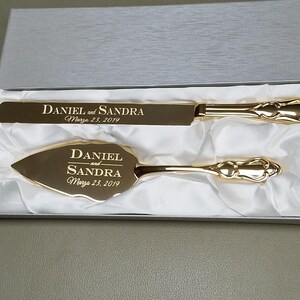 May include: Gold wedding cake knife and server set in a white box. The knife and server are engraved with the names "Daniel and Sandra" and the date "March 23, 2019".