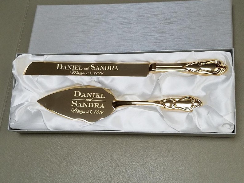 May include: Gold wedding cake knife and server set in a white box. The knife and server are engraved with the names "Daniel and Sandra" and the date "March 23, 2019".