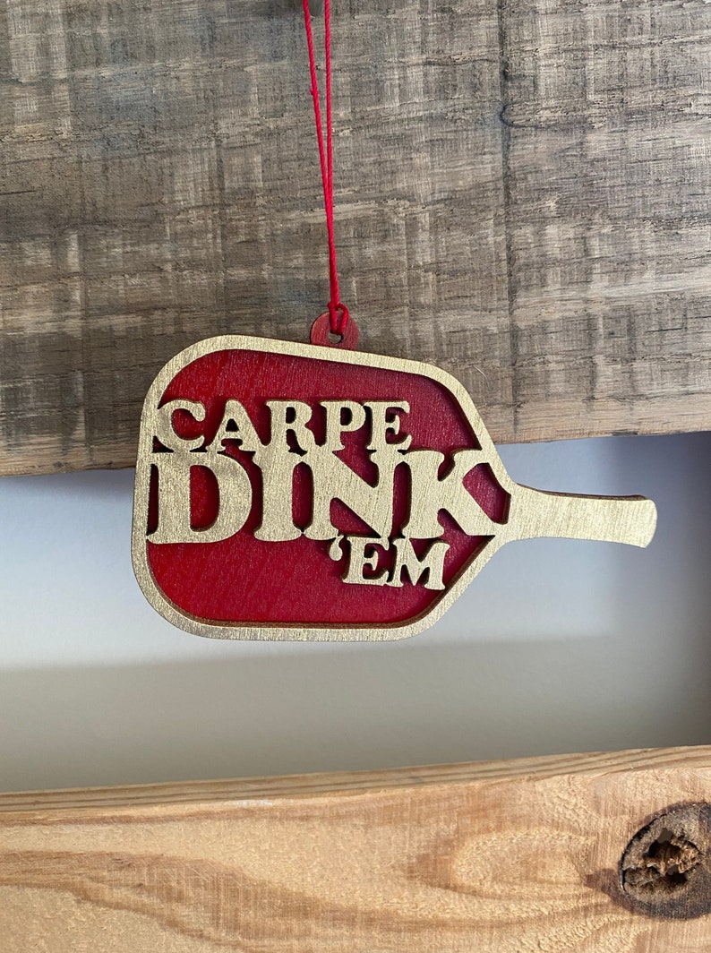 May include: A red wooden ornament shaped like a paddle ball with gold lettering that reads "CARPE DINK 'EM".