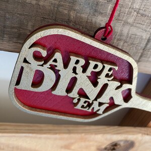 May include: A red and gold wooden ornament shaped like a paddle with the text "CARPE DINK 'EM" in gold lettering.