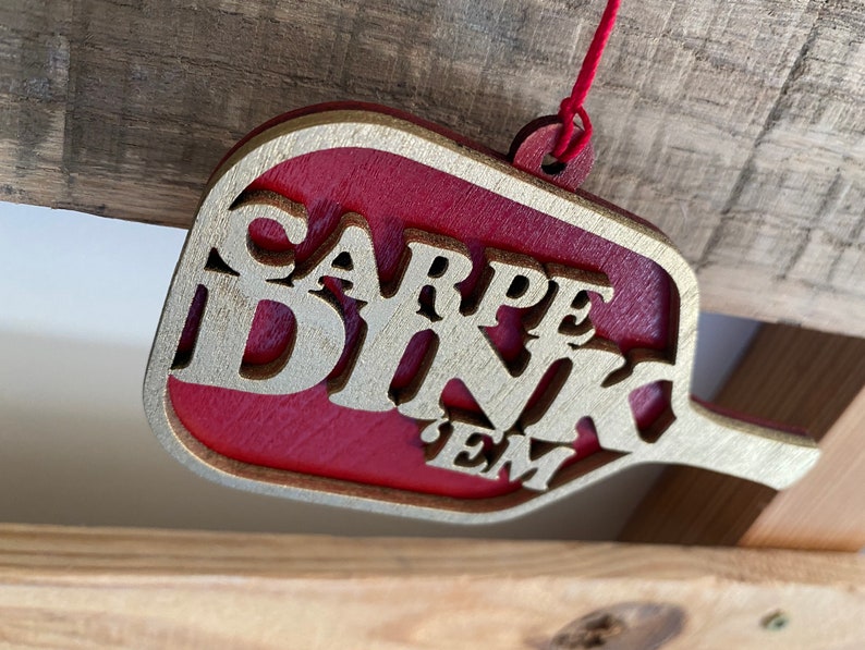 May include: A red and gold wooden ornament shaped like a paddle with the text "CARPE DINK 'EM" in gold lettering.