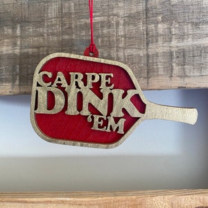 May include: A red wooden ornament shaped like a paddle ball with the words "CARPE DINK 'EM" in gold lettering.