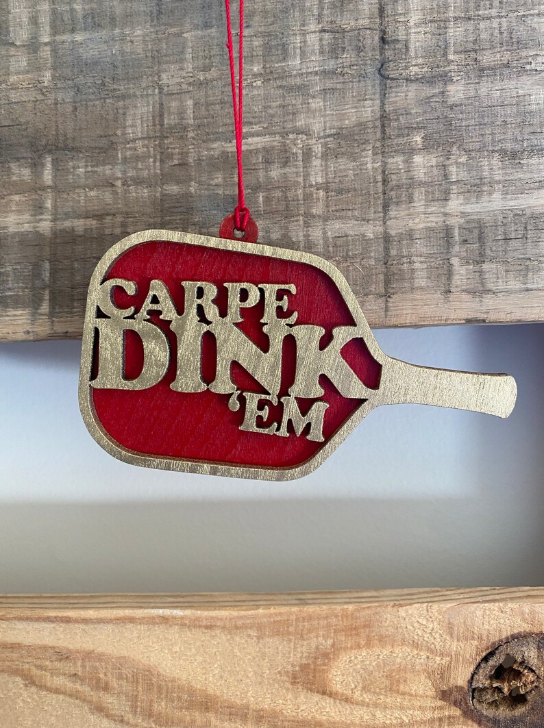 May include: A red wooden ornament shaped like a paddle ball with the words "CARPE DINK 'EM" in gold lettering.