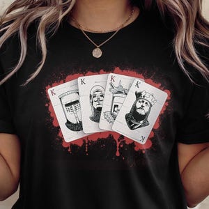 May include: Black t-shirt featuring a graphic of four playing cards with the King symbol and illustrations of historical figures. The cards are set against a red splatter design.