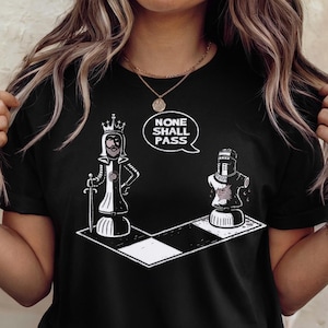 Puede incluir: Camiseta negra con un diseño de ajedrez. El diseño muestra dos piezas de ajedrez, una reina y un caballero, en un tablero de ajedrez, con el texto "NONE SHALL PASS" en un bocadillo. La reina sostiene una espada.