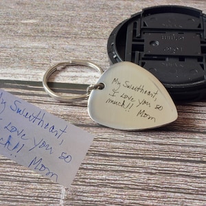 May include: A heart-shaped silver keychain with the handwritten message "My Sweetheart, I Love you so much!! Mom". A handwritten note with the same message is next to the keychain. A black object is in the background.
