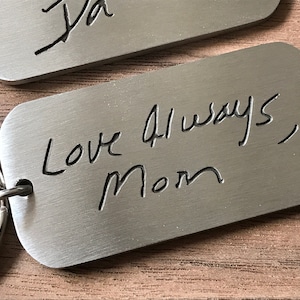 Könnte beinhalten: Zwei Schlüsselanhänger aus gebürstetem Metall mit handschriftlichen Inschriften. Einer lautet "Ja" und der andere "Love Always, Mom". Die Schlüsselanhänger haben abgerundete Ecken und sind an silbernen Schlüsselringen befestigt.