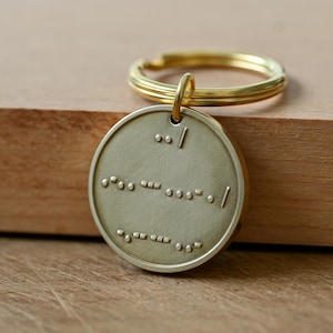 May include: A circular brass keychain featuring embossed Morse code patterns on its front surface. The metal disc is attached to a standard gold-toned split ring, providing a functional and metallic accessory with a distinct tactile design.
