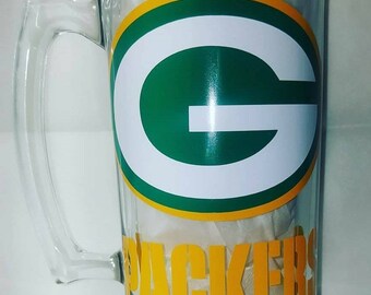 May include: A clear glass beer mug with a green and gold Green Bay Packers logo and the word "PACKERS" in yellow lettering.