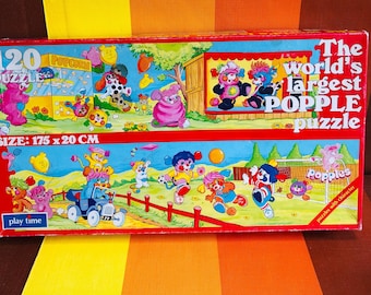 May include: A red and blue box for a 120 piece Popples puzzle. The box shows a cartoon image of the Popples characters playing in a field and at a popcorn stand. The box says "The world's largest Popple puzzle" and "puzzles with character". The puzzle measures 175 x 20 cm.