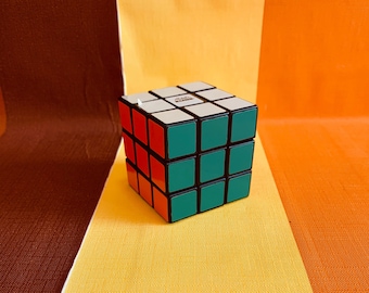 May include: A classic black, red, and green Rubik's Cube puzzle toy. The cube is made of plastic and has a white sticker on the top with the word "Rubik's" printed on it.
