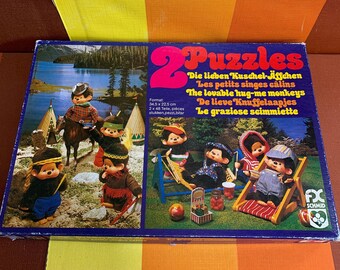 May include: Two puzzles featuring illustrations of the Hug-Me Monkeys. The first puzzle shows the monkeys in a Native American setting, riding a horse, and sitting in a teepee. The second puzzle shows the monkeys relaxing in a hammock and on a lounge chair. The box is blue with white text that reads "Die lieben Kuschel-Affchen", "Les petits singes câlins", "The lovable hug-me monkeys", "De lieve Knuffelaapjes", "Le graziose scimmiette", and "Format: 34,5 x 22,5 cm 2 x 48 Teile, pièces, stukken, pezzi, bitar".