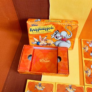 Könnte beinhalten: Eine Diddl Knuffldiffeis Spielbox mit einem leuchtend orangenen Hintergrund und einer Comicfigur auf der Vorderseite. Die Box ist geöffnet und die Spielsteine sind sichtbar. Die Spielsteine sind orange und zeigen Comicfiguren.