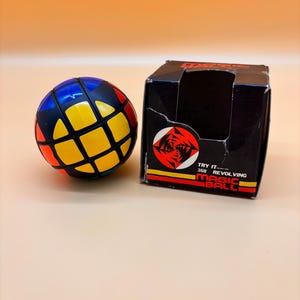 May include: A spherical puzzle toy with black grid lines and colored segments in yellow, blue, and orange. Beside it sits a black cardboard box featuring a circular red and white graphic, the text "TRY IT 360 REVOLVING MAGIC BALL," and yellow and red stripes.