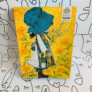 May include: A 500-piece Muva puzzle box featuring an illustration of a person in a large blue bonnet and patchwork dress standing in a field of yellow flowers. The box displays the text Muva Valkenburg (L) Puzzel 500 49 x 36 cm.