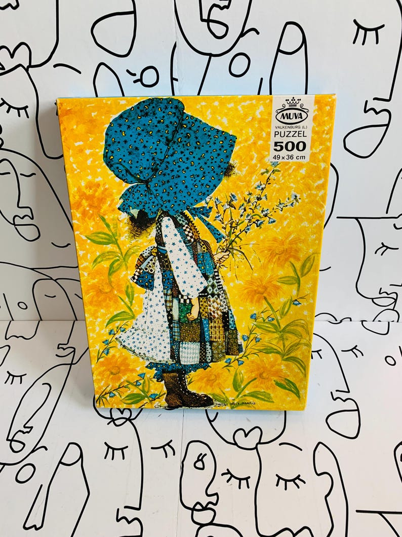 May include: A 500-piece Muva puzzle box featuring an illustration of a person in a large blue bonnet and patchwork dress standing in a field of yellow flowers. The box displays the text Muva Valkenburg (L) Puzzel 500 49 x 36 cm.
