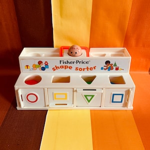 Może przedstawiać: Biała plastikowa zabawka Fisher-Price do sortowania kształtów z uśmiechniętą buzią dziecka na górze. Zabawka ma cztery otwory o różnych kształtach: koło, kwadrat, trójkąt i prostokąt. Każdy otwór ma odpowiadający mu kształt, który można włożyć do niego. Zabawka została zaprojektowana, aby dzieci uczyły się kształtów i kolorów.