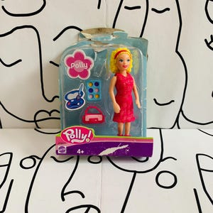 Może przedstawiać: Lalka Polly Pocket w różowej sukience, z różową torebką i niebiesko-różową paletą kosmetyków. Lalka jest zapakowana w przezroczystą plastikową skrzyneczkę z różowo-fioletowym tłem. Na pudełku widnieje napis "Polly Pocket" i "4+."