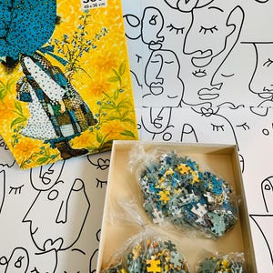 May include: A vintage 500-piece puzzle in a yellow box with a floral and figure design. The box measures 19.29 x 14.17 inches. Inside the box are bags of puzzle pieces in shades of yellow, blue, and green. The background is a white surface with black line art faces.