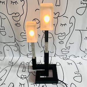 Può includere: Una moderna lampada da tavolo con due paralumi rettangolari in vetro smerigliato che emettono un bagliore caldo. Le lampade sono sostenute da steli in metallo nero e una base quadrata. Lo sfondo è un tessuto bianco con un disegno a linee nere ripetuto di volti.