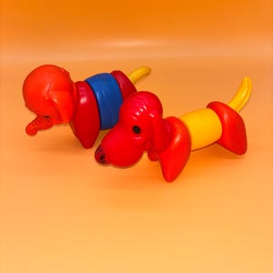 Può includere: Due giocattoli colorati a forma di bassotto. Uno è rosso, blu e giallo, l'altro è rosso e giallo. I giocattoli sono in plastica e hanno un design giocoso. Lo sfondo è arancione.