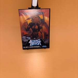 Può includere: Una cartuccia per videogiochi Sega Mega Drive per Altered Beast. La scatola del gioco presenta una bestia umanoide muscolosa con la testa di un leone. Il titolo "Altered Beast" è in bianco e blu. Lo sfondo è arancione acceso.