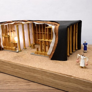 Op de afbeelding: Gedetailleerd model van een religieuze structuur met een zwart dak en goudkleurige pilaren. Miniatuurfiguren en een gouden kom zijn geplaatst op een zandachtige ondergrond, alles op een houten basis. Het interieur is verlicht.