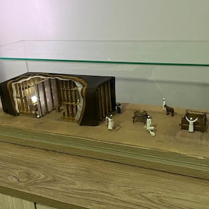 Op de afbeelding: Een miniatuur diorama van een bijbelse scène in een glazen vitrine. Het diorama bevat een houten structuur met een licht erin, figuren van mensen en een klein dier. Het diorama staat op een zandbasis.