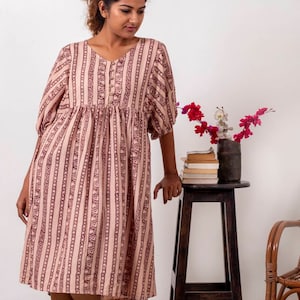 Peut inclure: Une femme portant une robe à rayures rose et marron avec un imprimé floral. La robe a un col en V et des manches 3/4. Elle se tient devant un mur blanc et un tabouret en bois avec un vase de fleurs dessus.