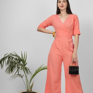 May include: A coral-coloured jumpsuit with a V-neck and wide legs. The jumpsuit has short sleeves and a wrap-style bodice. A small black handbag with gold-coloured hardware is held in the hand. A yellow bracelet is worn on the wrist.