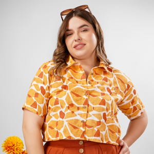 Könnte beinhalten: Eine Frau trägt ein kurzärmeliges, gelb-orangefarbenes, gemustertes Hemd mit Knöpfen und einem verkürzten Saum. Das Hemd hat einen weißen Hintergrund mit einem geometrischen Muster.