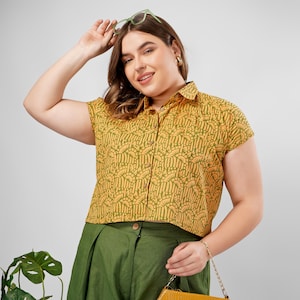 Op de afbeelding: Een shirt met korte mouwen met een geel en groen patroon, gecombineerd met een groene broek. Het shirt heeft een kraag en een herhalend ontwerp. De outfit is geaccessoireerd met een gouden armband en een gele handtas.