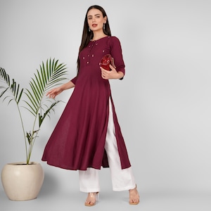 Könnte beinhalten: Eine lange, dunkelrote Kurta mit einem weißen Seitenschlitz und einem Knopfverschluss. Die Kurta hat ein dezentes Stickmuster am Ausschnitt. Das Model trägt eine weiße Hose und hält eine kleine rote Clutch.