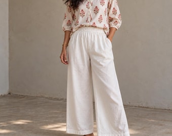 Puede incluir: Una blusa de color crema con estampado floral en rojo y verde, mangas tres cuartos y cierre de botones frontal, combinada con pantalones anchos de color blanco roto con cintura elástica.