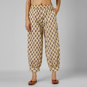 May include: Cream-coloured, floral-print trousers with a relaxed fit. The trousers feature a repeating pattern of purple flowers and green leaves. The elastic waistband and cuffed ankles create a comfortable, casual style. The trousers are made of a lightweight fabric.