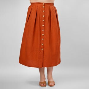 Puede incluir: Una falda midi de color óxido con botones en la parte delantera y pliegues. La falda está hecha de una tela similar al lino.