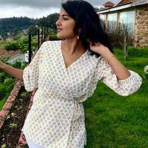 Pode incluir: Um top branco estilo wrap com estampa floral amarela e um laço na cintura. O top tem mangas 3/4 e decote em V.