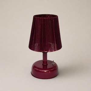 May include: A small, burgundy-colored table lamp with a pleated velvet lampshade. The lamp has a round base, a slender stem, and a small on/off switch. The shade is a deep red-purple color, and the base and stem have a glossy finish.