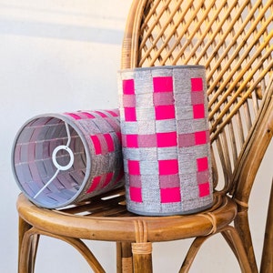 May include: Two grey and pink gingham lampshades sit on a wicker chair. The lampshades have a woven texture and a pink and grey gingham pattern.