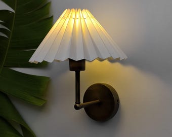 May include: A wall-mounted lamp with a white, pleated lampshade and a bronze-colored metal base. The lamp is lit, casting a warm glow. The design is simple and elegant, suitable for various interior styles.