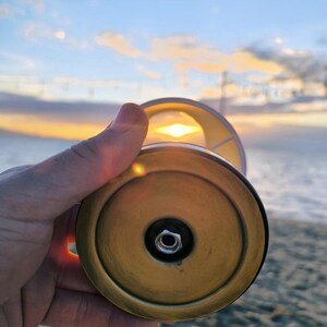 May include: A brass-colored lamp base with a white lampshade is held up against a sunset. The lamp base has a circular design with a central nut. The background shows a beach and the sky with the sun setting.