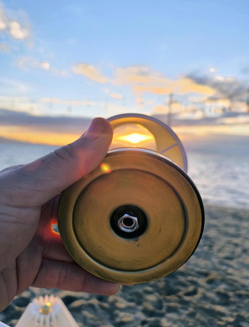 May include: A brass-colored lamp base with a white lampshade is held up against a sunset. The lamp base has a circular design with a central nut. The background shows a beach and the sky with the sun setting.